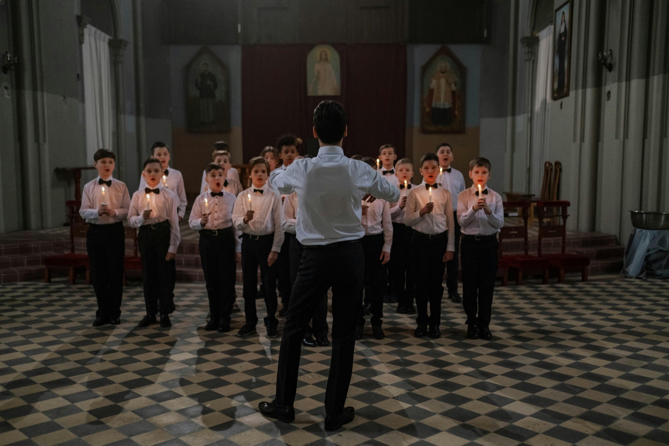 A boys choir dressed in uniforms singing in a candlelit church ceremony led by a conductor.